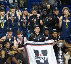 A shot of the University of Victoria men's basketball team wearing navy blue jerseys with gold medals and a U Sports championship banner.
