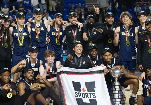 A shot of the University of Victoria men's basketball team wearing navy blue jerseys with gold medals and a U Sports championship banner.