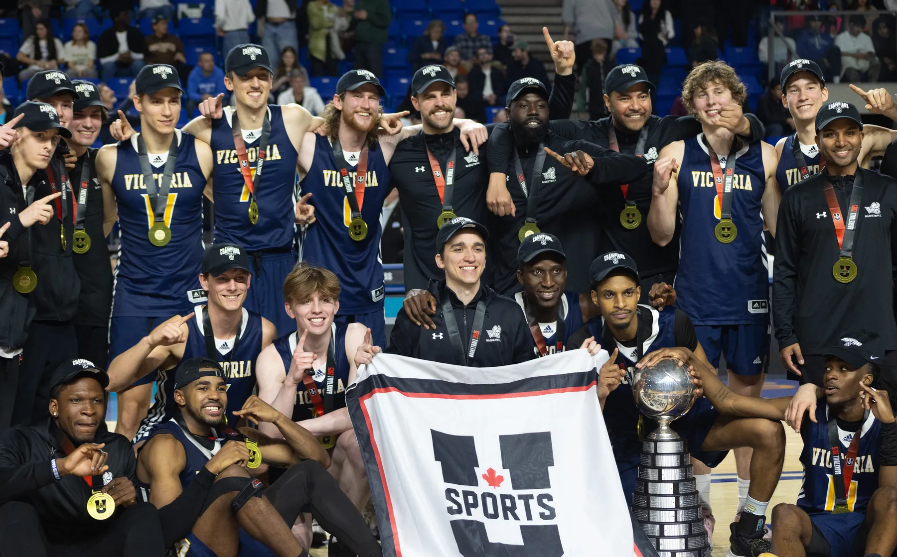 A shot of the University of Victoria men's basketball team wearing navy blue jerseys with gold medals and a U Sports championship banner.
