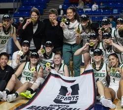 Woman basketball players in white jersey kits sit around a U Sports banner wearing black "national champions" hats.