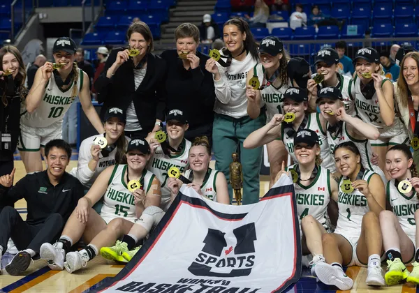 Woman basketball players in white jersey kits sit around a U Sports banner wearing black "national champions" hats.