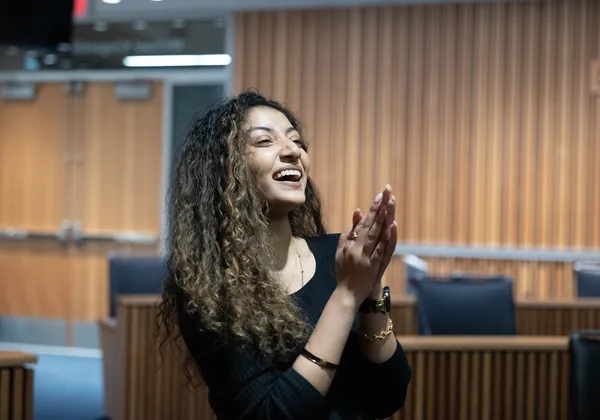 A photo of a woman in a black dress, smiling with her hands clapped together.