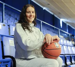A headshot of Olivia Weekes sitting in the stands of War Memorial Gym with a basketball in her lap.