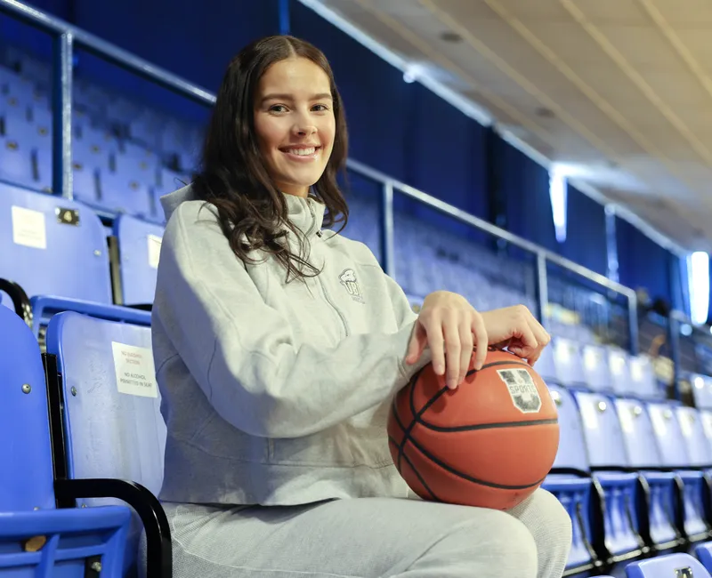 A headshot of Olivia Weekes sitting in the stands of War Memorial Gym with a basketball in her lap.