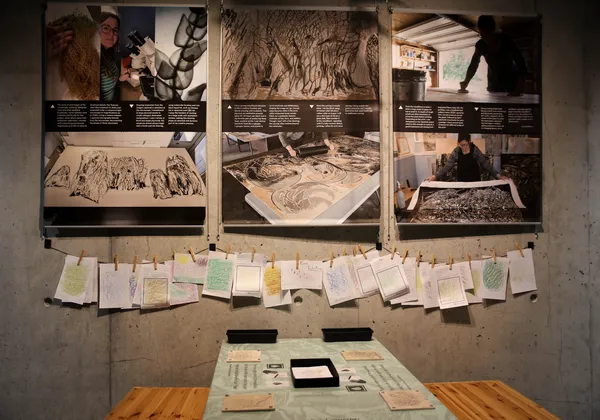 Three panels with text and images of Matthias making prints. Sheets of paper are hung on twine beneath them.