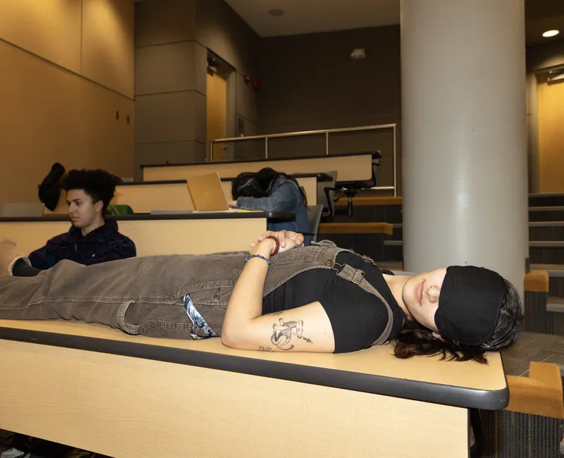A student wearing a sleeping mask and lying across a desk in a lecture hall.