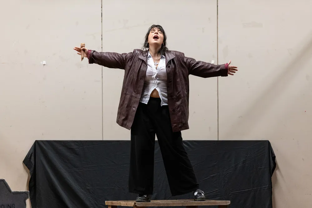 A white woman in a leather jacket stands and sings on a stage
