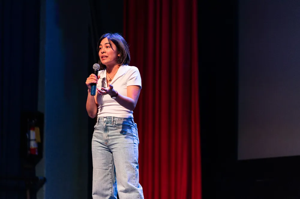 Kat Jayme, a woman in jeans and a white T-shirt, stands on a stage speaking into a microphone.