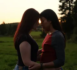 Two girls press their foreheads together, wrapping their arms around the other's waist. They stand in a green field, and the sun sets in the background.