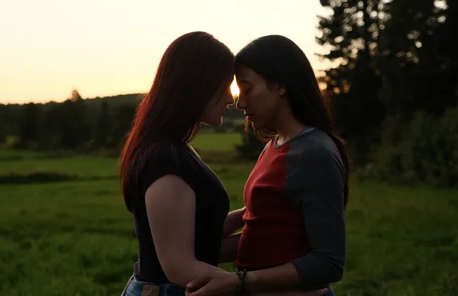 Two girls press their foreheads together, wrapping their arms around the other's waist. They stand in a green field, and the sun sets in the background.