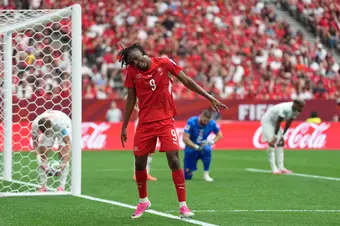 Manzambi dances to celebrate in front of Canada's net, while Canada's back line crouches over, distraught.