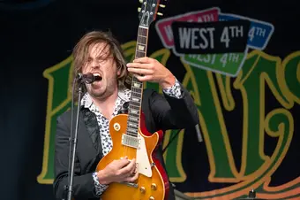 A man shreds a guitar with a Khatsahlano sign in the background.