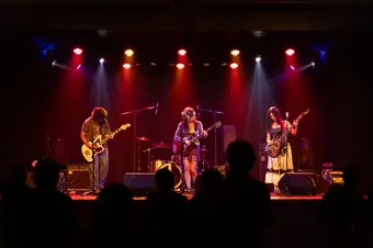 3 members of a band play instruments on a stage, with a small crowd