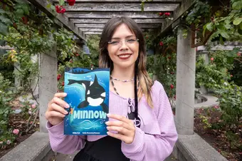 A woman smiles in a garden, holding a graphic novel with a whale on the cover.