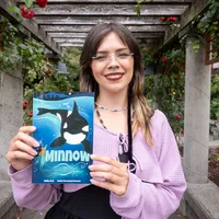 A woman smiles in a garden, holding a graphic novel with a whale on the cover.