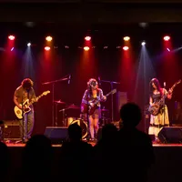 3 members of a band play instruments on a stage, with a small crowd