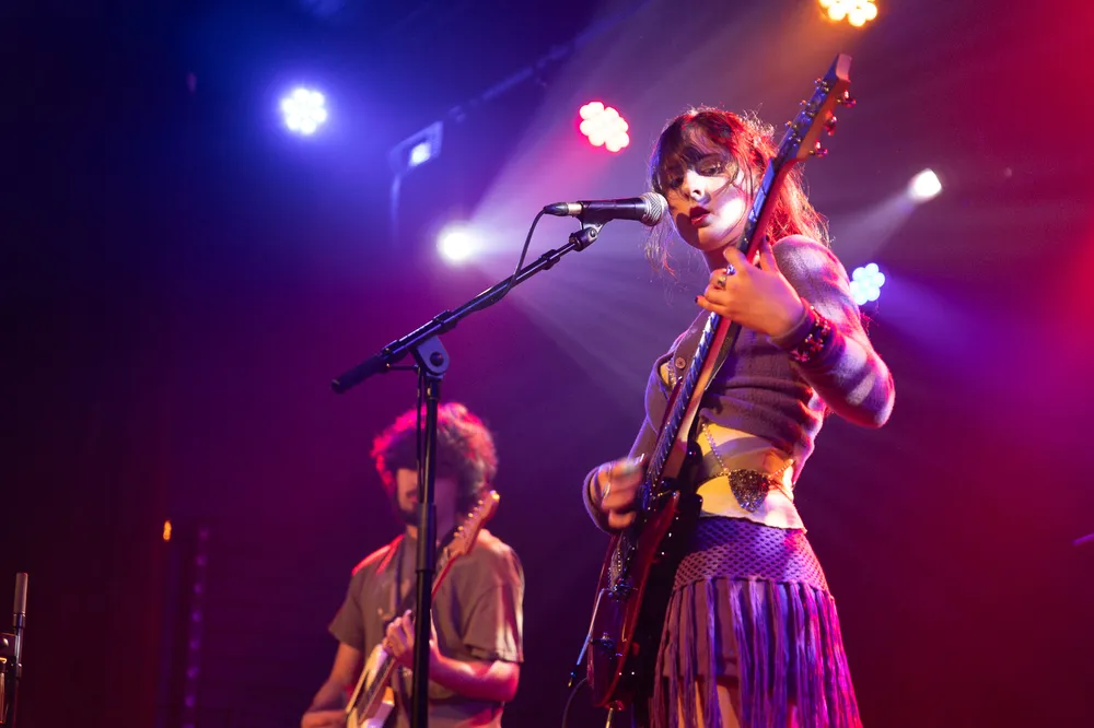 A female guitarist wearing purple stands at a microphone