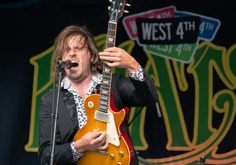 A man shreds a guitar with a Khatsahlano sign in the background.