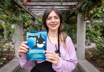 A woman smiles in a garden, holding a graphic novel with a whale on the cover.