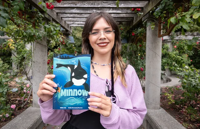 A woman smiles in a garden, holding a graphic novel with a whale on the cover.