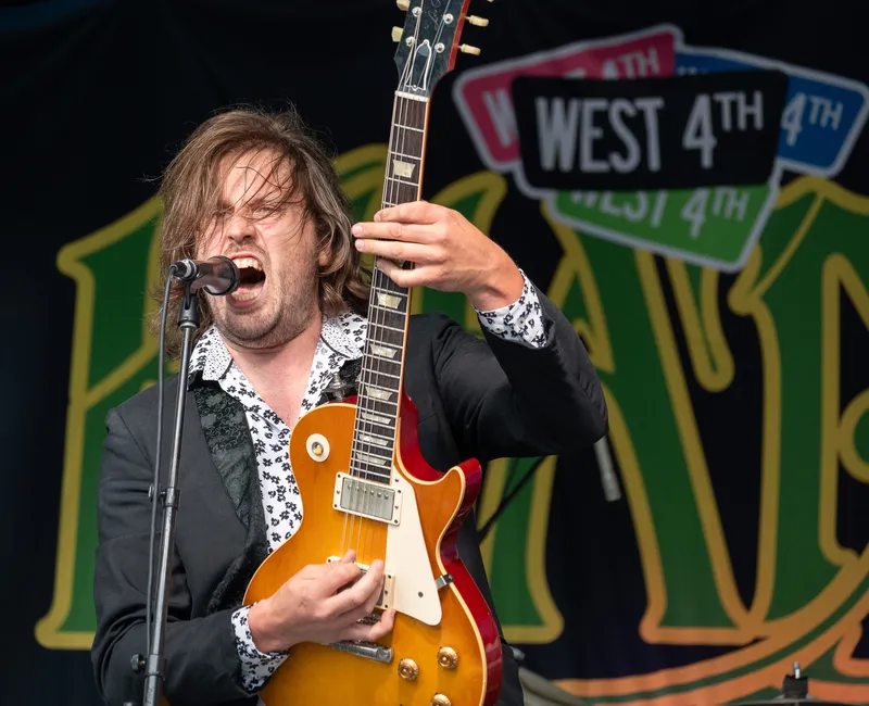 A man shreds a guitar with a Khatsahlano sign in the background.