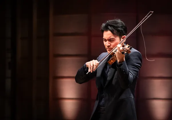 A man with black hair plays a violin onstage. He is dressed in black.