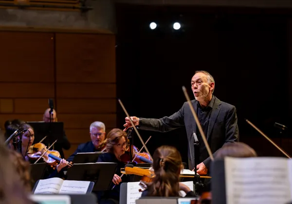 A conductor of an orchestra stands in front of violinists. He is wearing a black suit and his arm is in mid-stroke.
