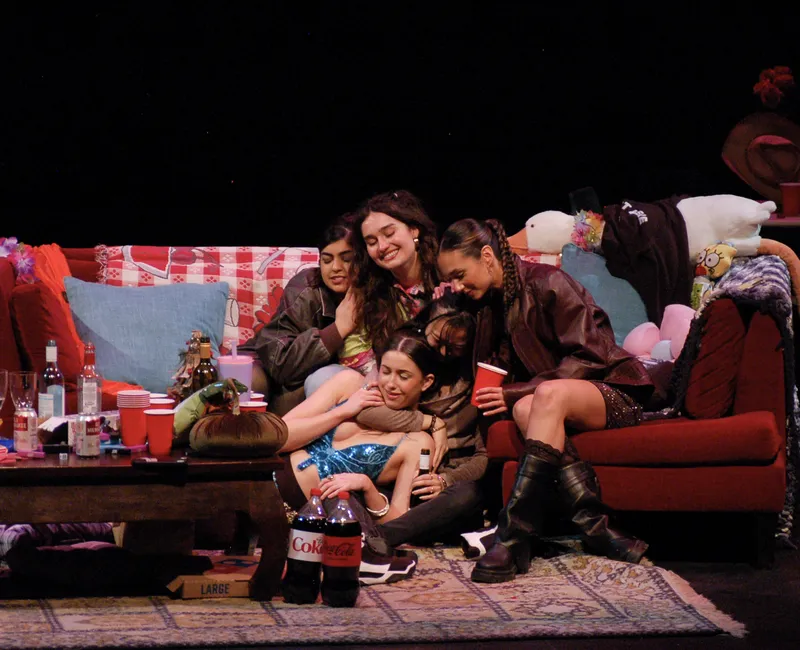 A group of women in their early '20s sit on a red couch, embracing each other. Bottles of alcohol, pop and plastic cups sit on a cluttered coffee table in front of them.