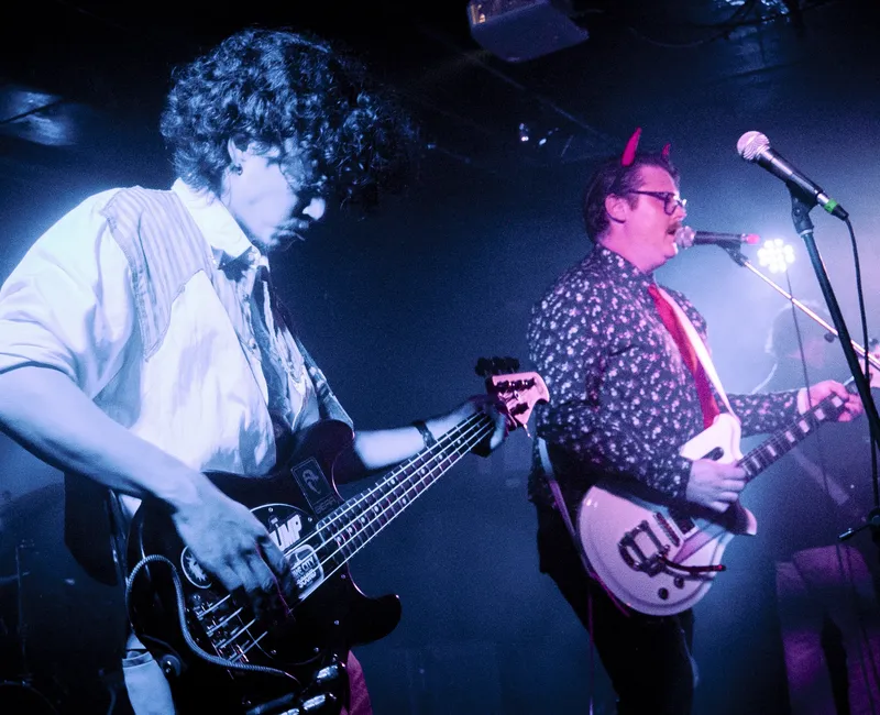Two people on stage in coloured lighting, one plays a bass and the other plays guitar while singing into a mic
