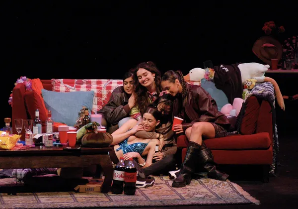 A group of women in their early '20s sit on a red couch, embracing each other. Bottles of alcohol, pop and plastic cups sit on a cluttered coffee table in front of them.