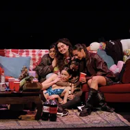 A group of women in their early '20s sit on a red couch, embracing each other. Bottles of alcohol, pop and plastic cups sit on a cluttered coffee table in front of them.
