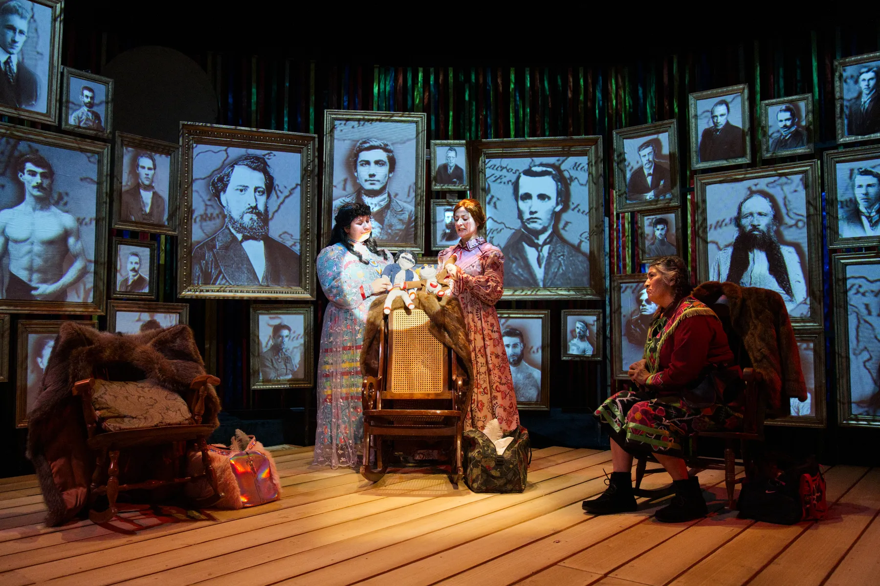 Two women stand before a wall of black and white portraits, while another one sits in a chair nearby.