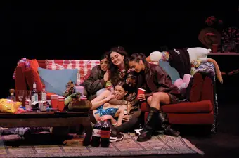 A group of women in their early '20s sit on a red couch, embracing each other. Bottles of alcohol, pop and plastic cups sit on a cluttered coffee table in front of them.