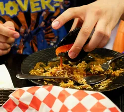 A mostly-eaten plate of yellow rice in a black table, with a hand hovering over it pouring a red sauce.