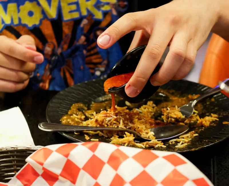 A mostly-eaten plate of yellow rice in a black table, with a hand hovering over it pouring a red sauce.