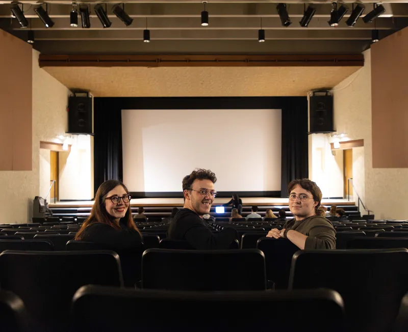 Three people sitting in a theatre, turned away from the screen and towards the camera