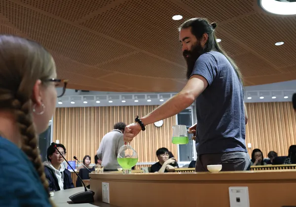 A standing man with a beard pours bright green tea into a cup.
