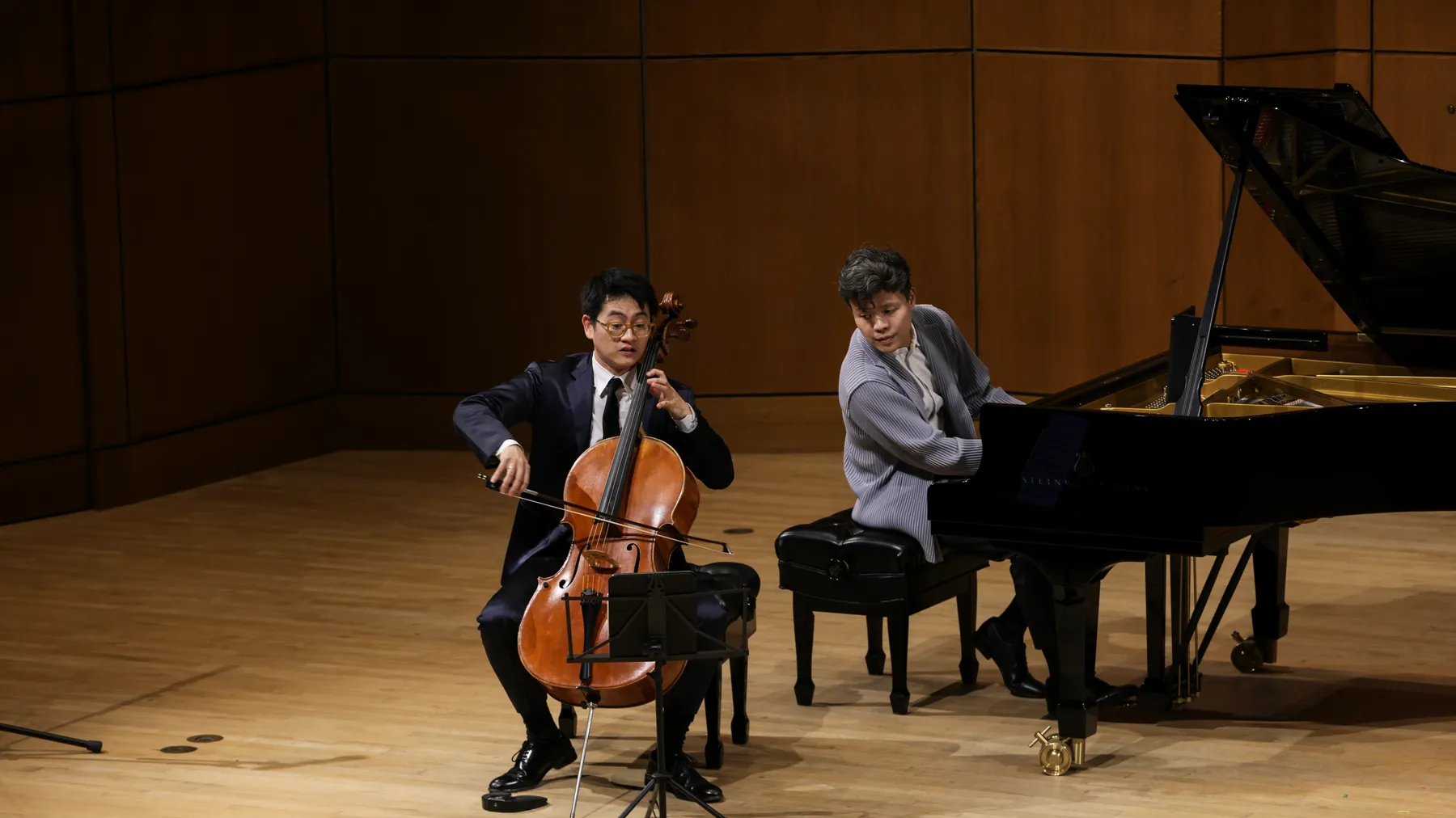 Two men sit on a stage, one playing a piano, the other playing a cello