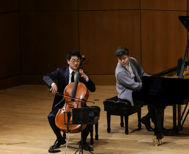 Two men sit on a stage, one playing a piano, the other playing a cello