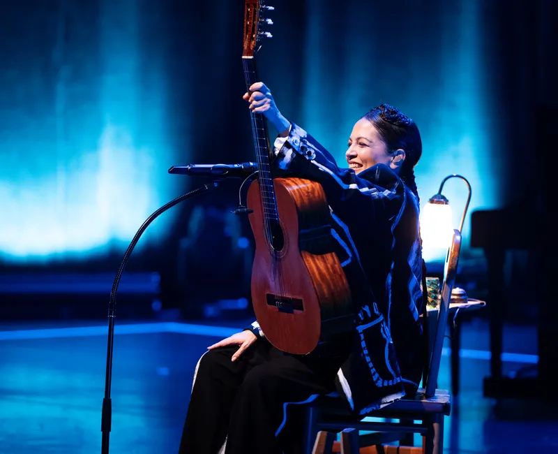 A woman on stage holds an acoustic guitar aloft