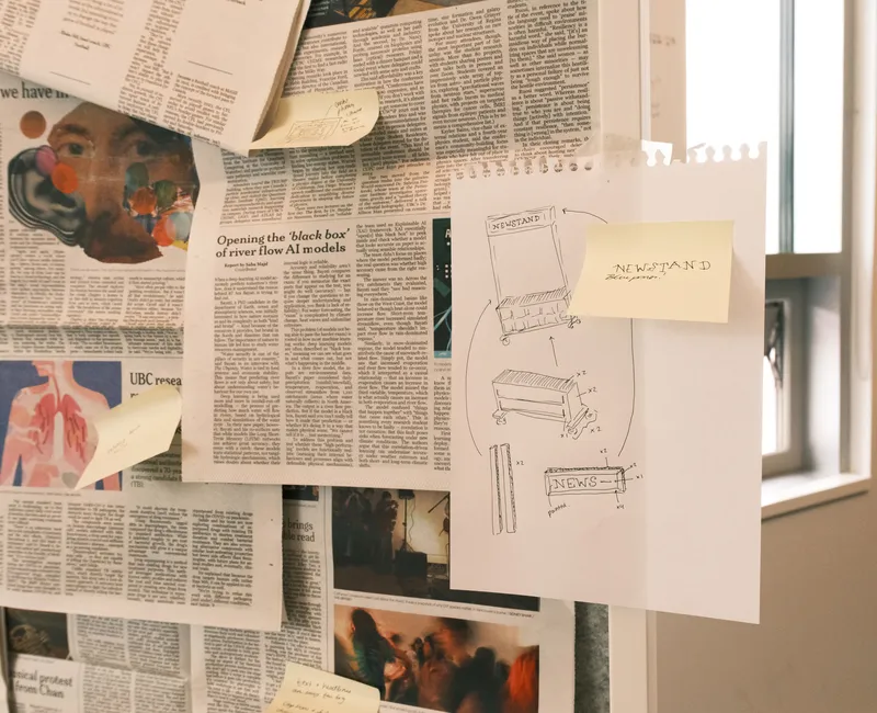 Newspaper clippings pinned up on a board. There are sticky notes stuck on the clippings with annotations. On the board, there's also a sheet of paper with the blueprint of a newsstand hand-drawn on it.
