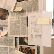 Newspaper clippings pinned up on a board. There are sticky notes stuck on the clippings with annotations. On the board, there's also a sheet of paper with the blueprint of a newsstand hand-drawn on it.