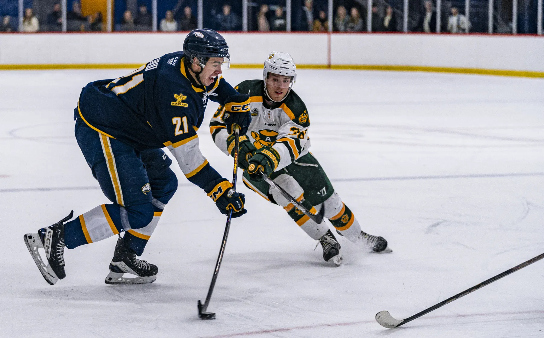 A hockey player in a navy blue jersey kit and helmet are on the left hand side of the image, getting ready to shoot the puck. Another player in a white jersey kit and white helmet in centered, facing the camera, attempting to stop the shot. They are both on an indoor ice rink with white boards. Some fans are blurry in the background, stadning by the boards.
