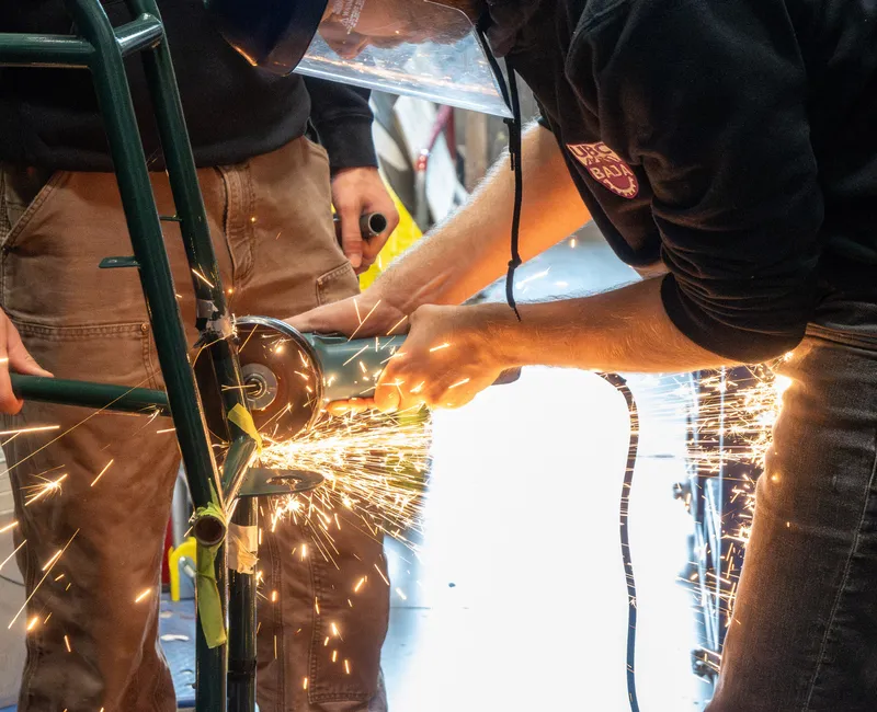 A person wearing a face shield takes a power tool to a metal frame, creating a spray of orange sparks.