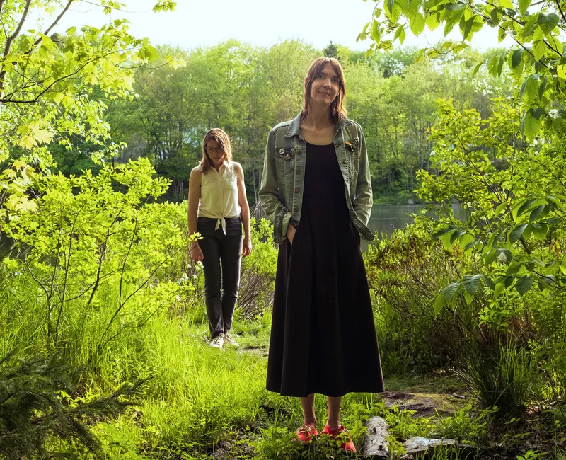 Carla Gillis, a white woman with brunette, shoulder-length hair, stands in the woods wearing a black maxi dress. Her sister Lynette, a white woman with glasses wearing a white top and black skinny jeans, stands behind her, looking at the ground.