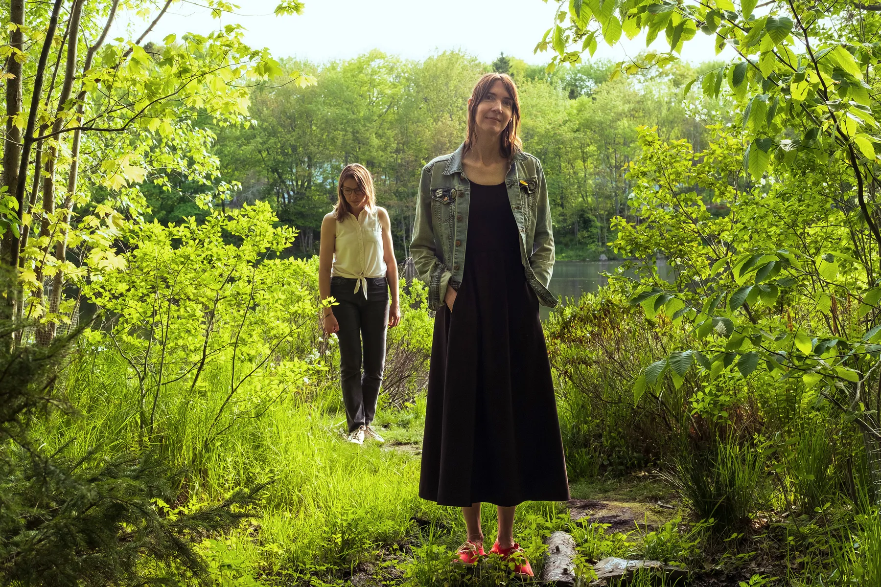 Carla Gillis, a white woman with brunette, shoulder-length hair, stands in the woods wearing a black maxi dress. Her sister Lynette, a white woman with glasses wearing a white top and black skinny jeans, stands behind her, looking at the ground.