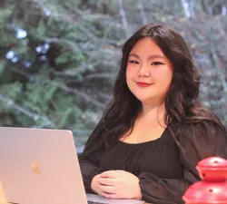 Emily Tang, a Chinese woman with long black hair and a black blouse, smiles while sitting at a table with a laptop in front of her.
