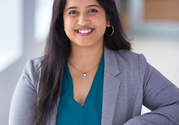 Rashmi Prakash, a smiling woman with long, dark hair, stands against a blurred background. She's wearing a teal blouse and light grey blazer.