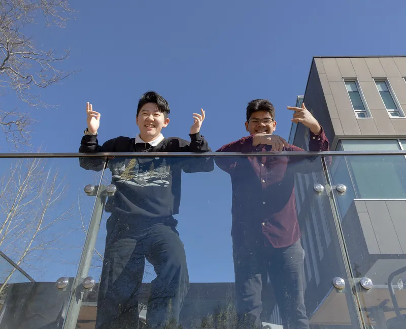Justina He wears a black sweatshirt and jeans and Nayis Majumder wears a burgundy button-down and jeans. Both lean over a glass barrier, smiling and throwing up peace signs.