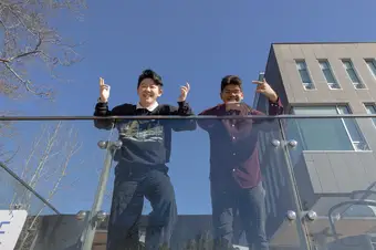 Justina He wears a black sweatshirt and jeans and Nayis Majumder wears a burgundy button-down and jeans. Both lean over a glass barrier, smiling and throwing up peace signs.